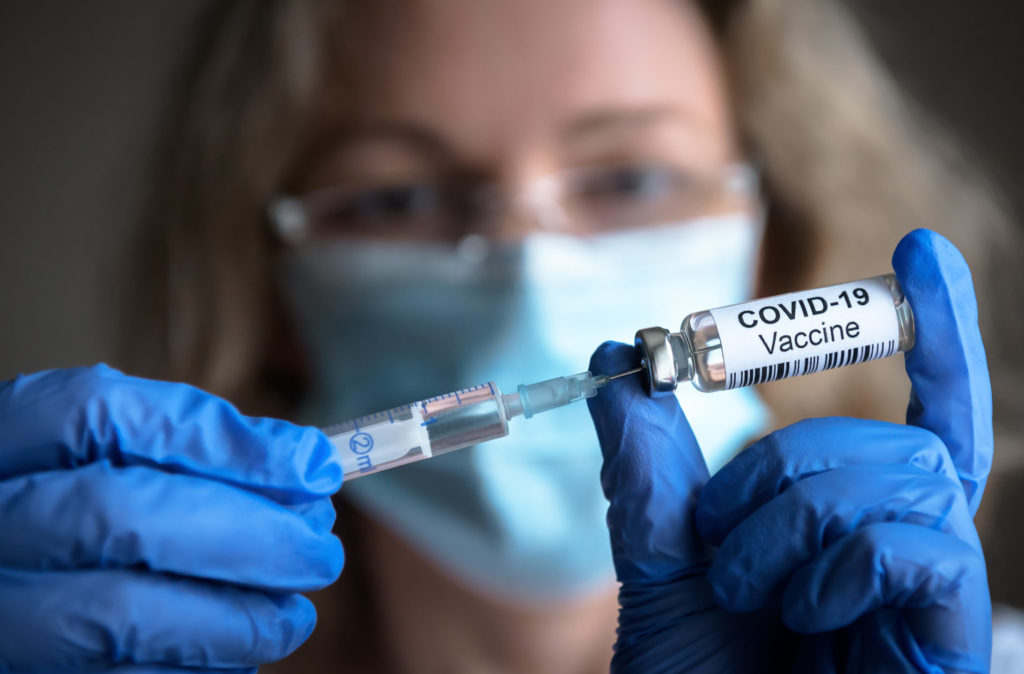 a female medic is holding a syringe stuck in a vile that has a covid-19 vaccine label on it